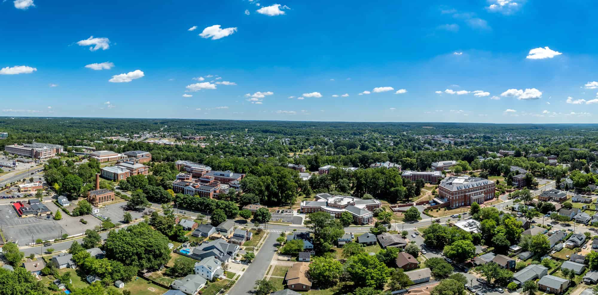 An aerial panorama of a tree-lined town with brick institutional buildings and surrounding residential streets under a blue sky with scattered clouds
