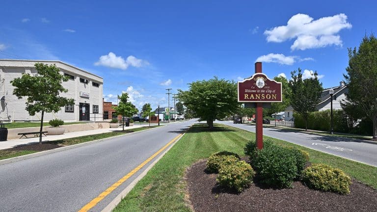 A road with a maroon welcome to Ranson city sign, a gray building, and scattered trees under a blue sky.