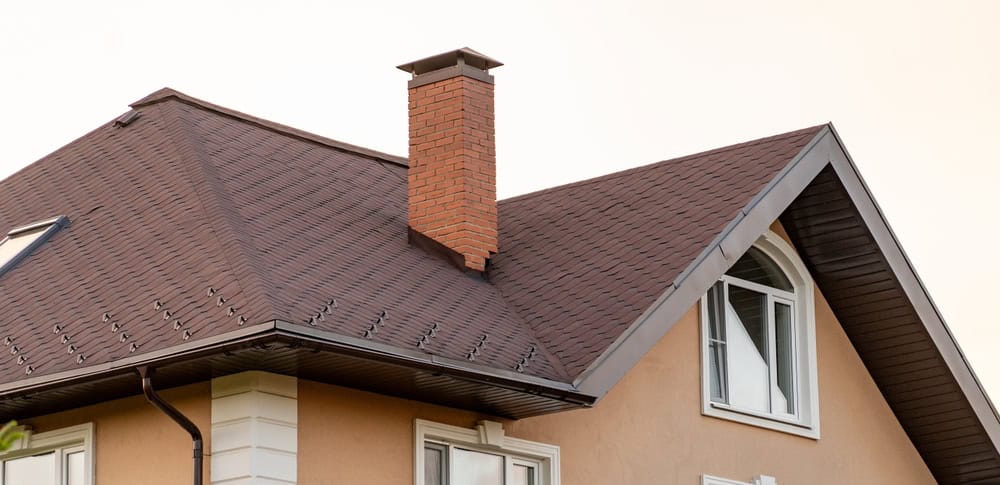 A modern house features a brown shingle roof, prominent brick chimney, and arched window under a soft evening sky.