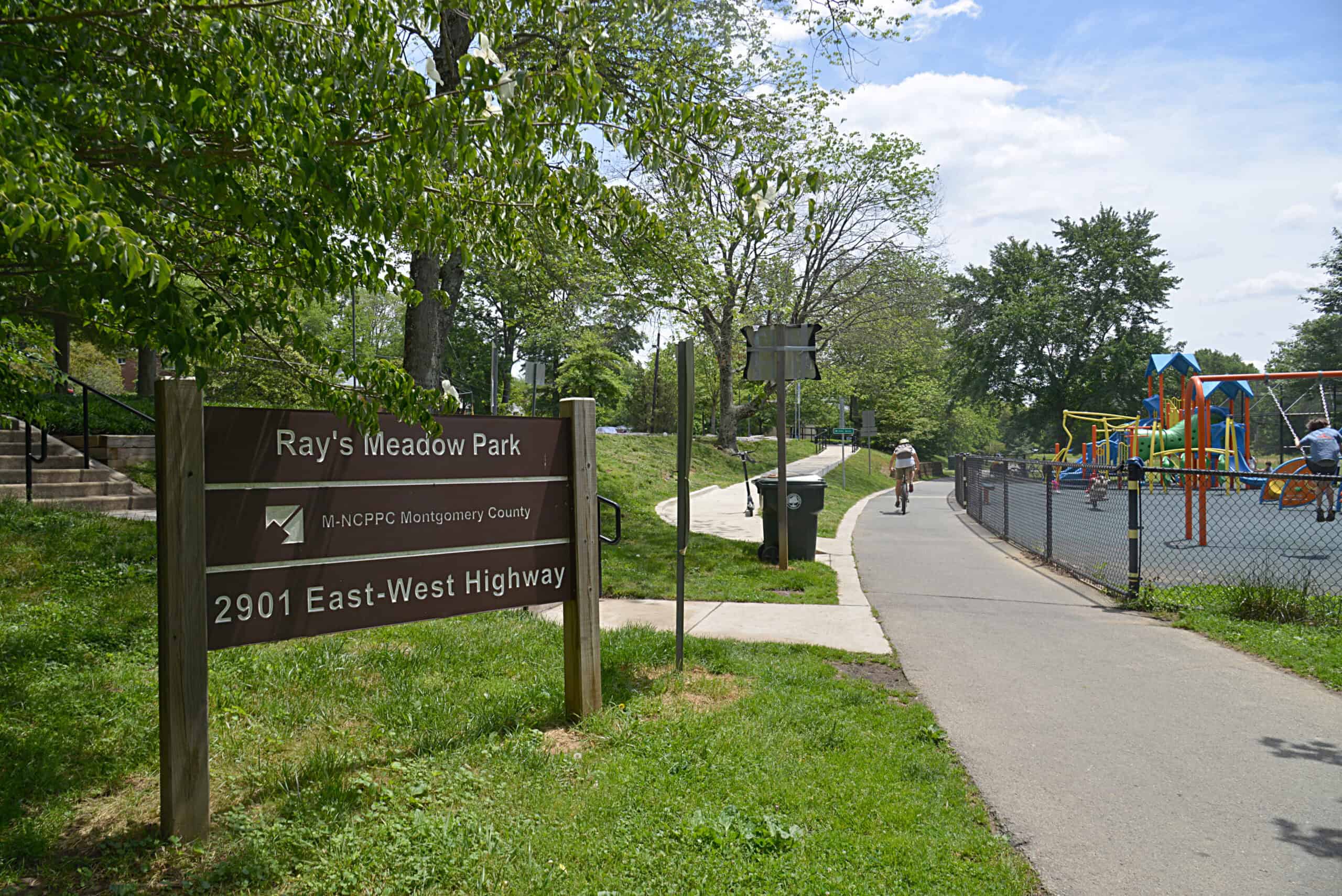Park sign, walking trail and colorful playground amid green trees at Ray’s Meadow Park in Chevy Chase.