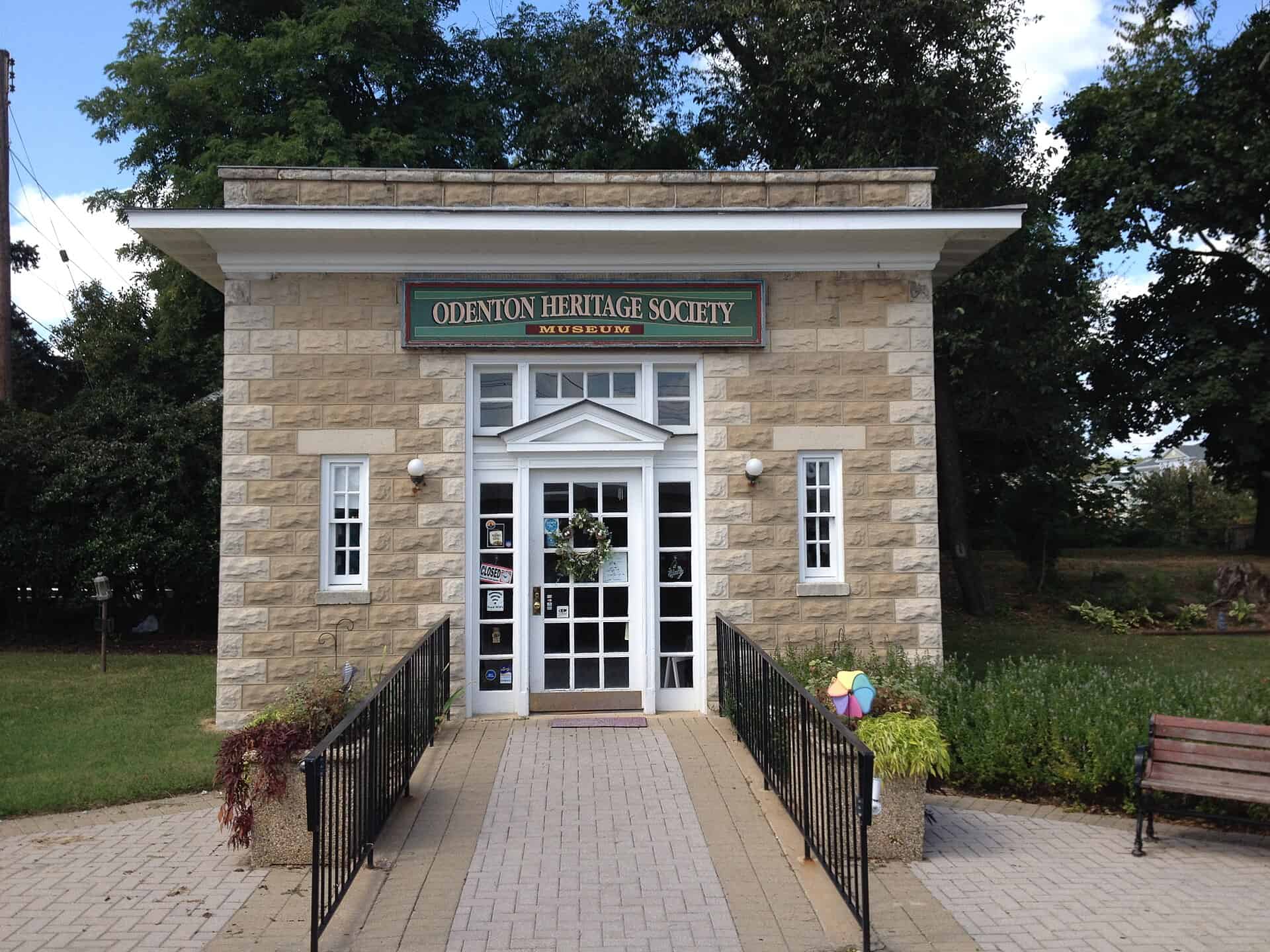 Odenton Heritage Society Museum entrance with a stone façade, landscaped walkway and leafy trees.