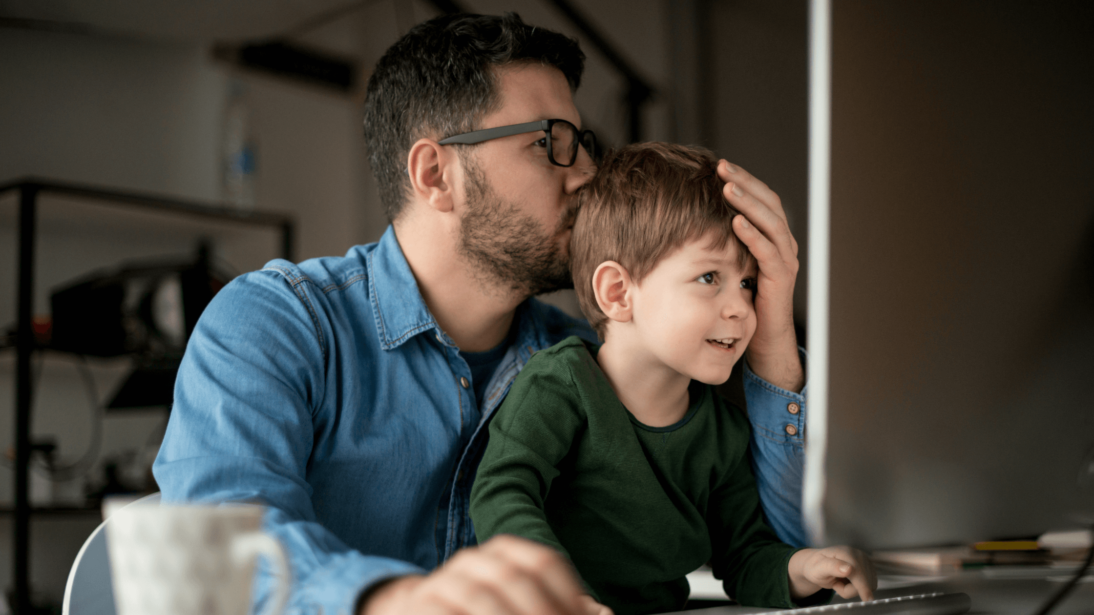 A father in glasses and a denim shirt gently kisses his young son on the head while they sit together in front of a computer, symbolizing a family business and the need to protect assets during a Texas divorce.