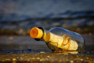 Clear bottle with a cork. Inside the bottle is a rolled up piece of paper with a string wrapped around it. The bottle is on a wet beach.