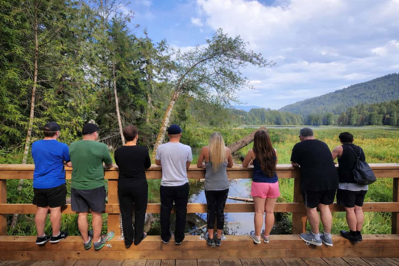 Group of men and women standing on a wodden bridge looking over a stream that goes through a wooded field to a lake in the distance.