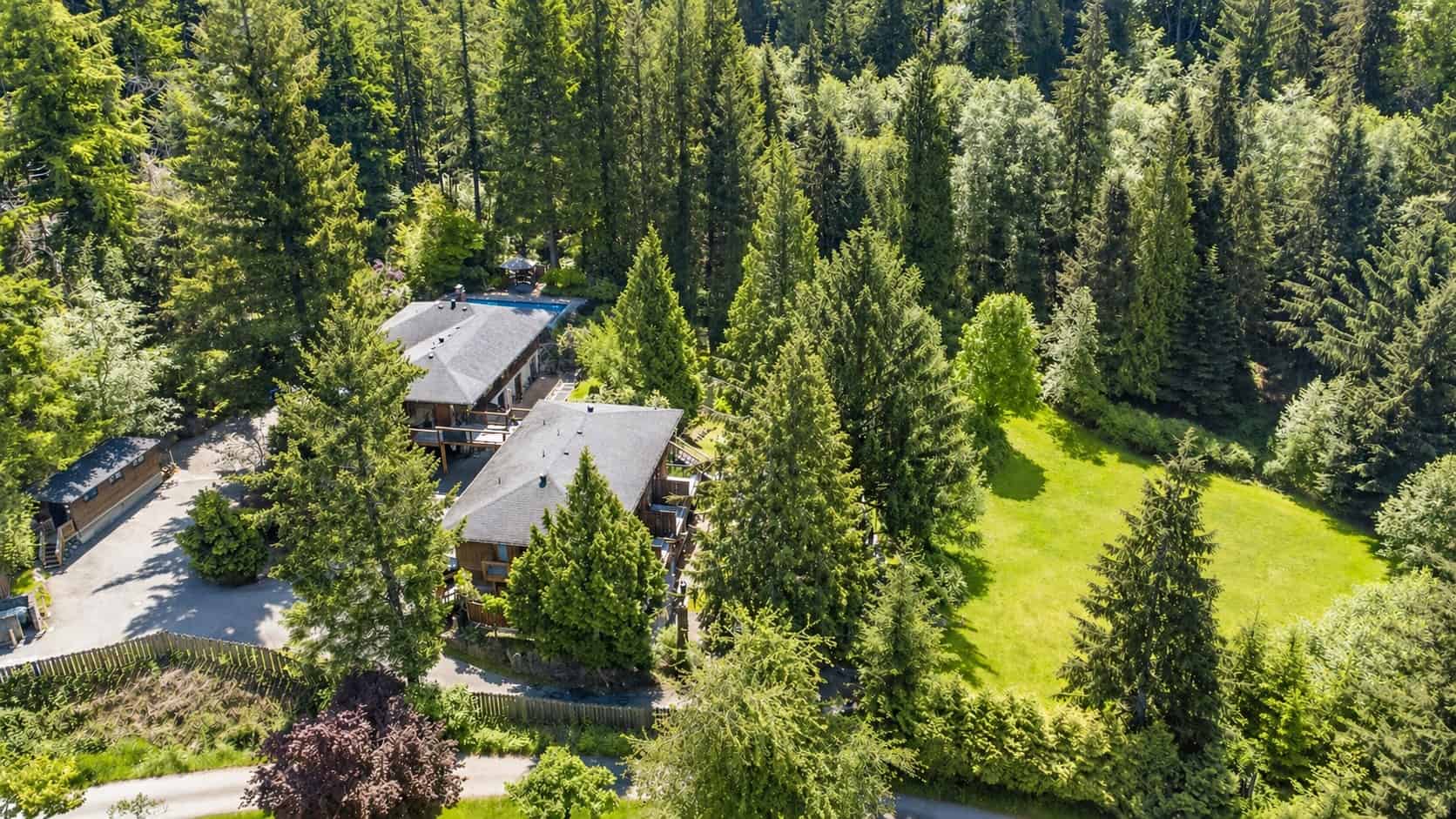 Two large wooden buildings with swimming pool. On the left side is a driveway with a smaller wooden building. The buildings are surrounded by large trees with a field