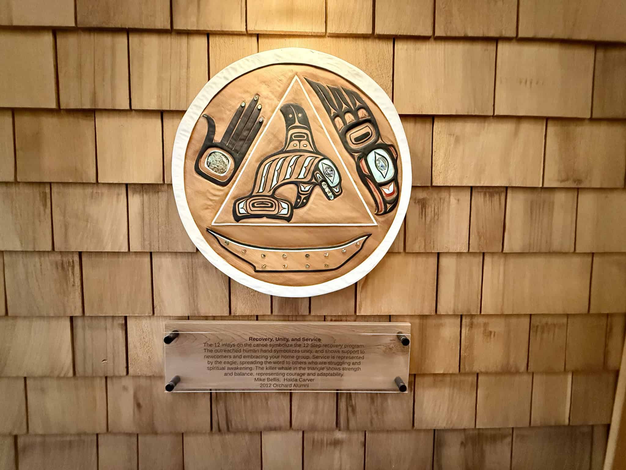Circular Haida carving with a triangle in the middle symbolizing Recovery, Unity and Service. Below the art is explaination of the symbols. 12 inlays on a canoe symbolizes the 12 Step recovery program. The ousreached hand symbolizes unity. Service is represented by an eagle. A killer whale in the center of the triangle shows strength and balance. Carved by Mike Bellis Haida Carver and 2012 Orchard Alumni.