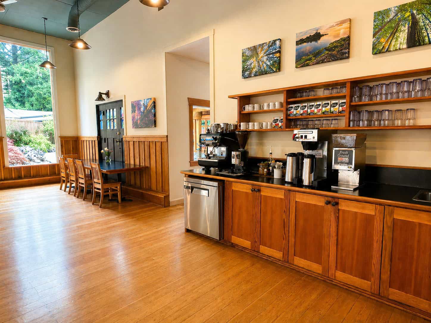Large counter with expresso machine, coffee/water brewer, coffee grider, many boes of different teas, coffee cups and glasses. A small aluminum fridge is below the expresso machine. A table with chairs is in the far side of the room. The floor is beautiful wood. THrough the windows a garden with flowers and trees can be seen.