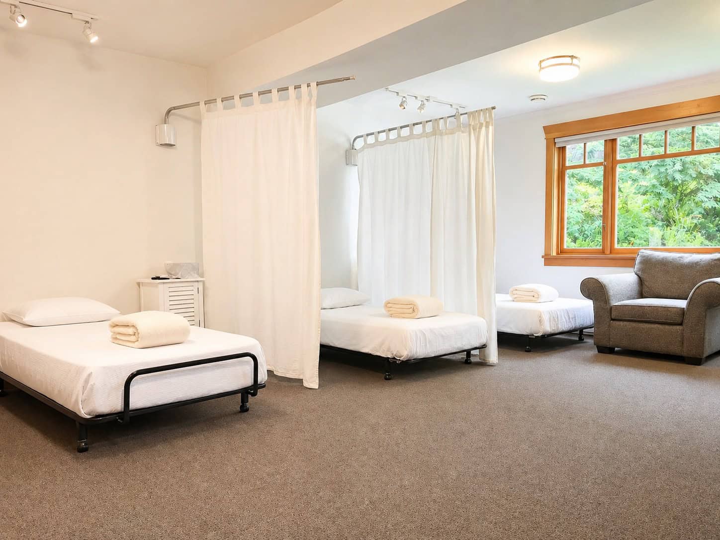 Large room with 3 beds and a comfortable chair in front of a large window with trees in the background. Pillows and blankets are on the beds with privacy curtains between them. The healing room at Orchard Recovery Center.
