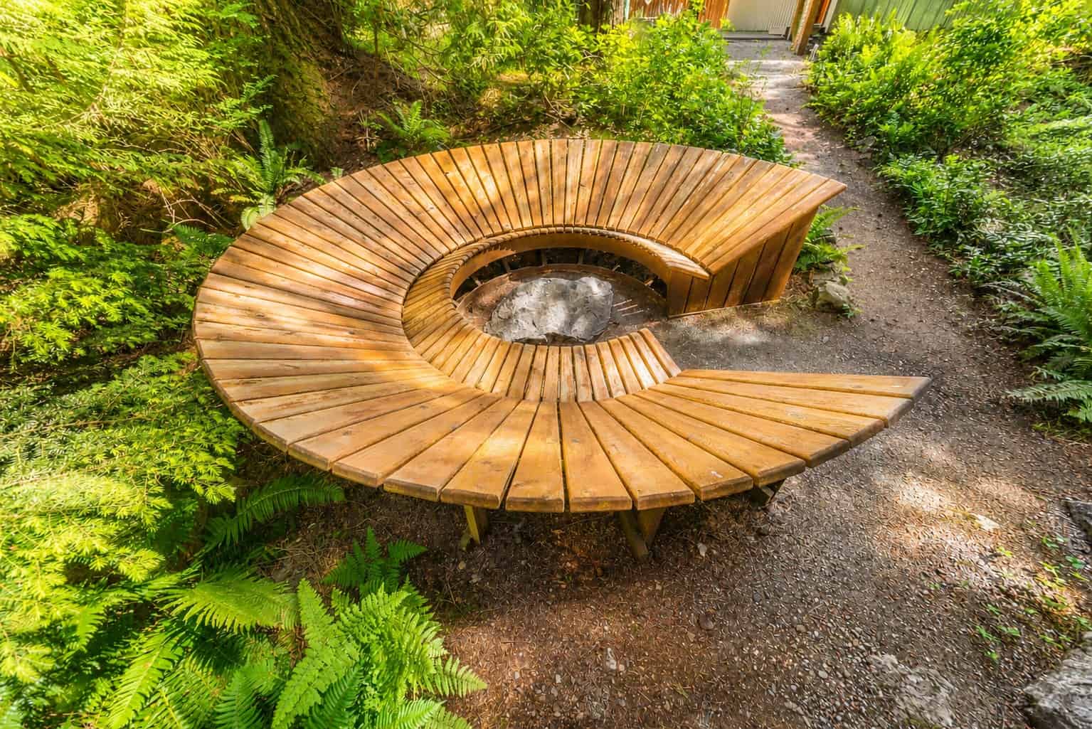 Wooden bench surrounding a large rock so you can lean back with your feet on the rock. Ferns, trees and green plants surround the bench. A gravel path goes past the bench.