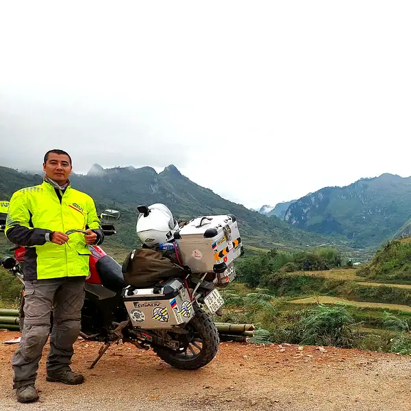 Rider beside a loaded touring motorcycle above a terraced valley on the Ha Giang Loop — Vietnam motorbike tours, Outriders Co.
