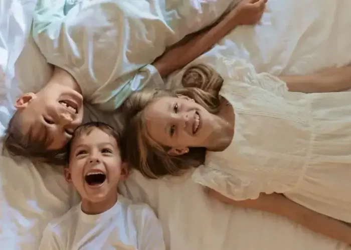 Family And Kids β Homes For Sale In Palmilla Brightly Lit Family Of Three Laughing And Lying On Bed In Palmilla Home, Showcasing Comfortable And Cozy Atmosphere For Palmilla Real Estate Listings.