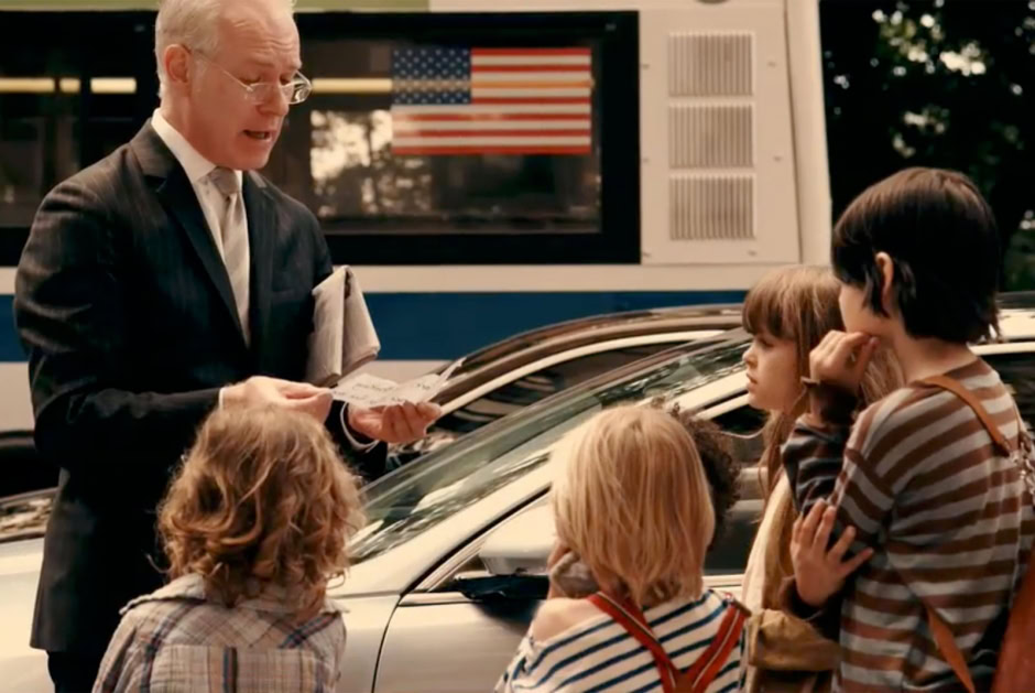 A man in a suit and glasses reads a newspaper to children on a city street with a bus and American flag in the background.