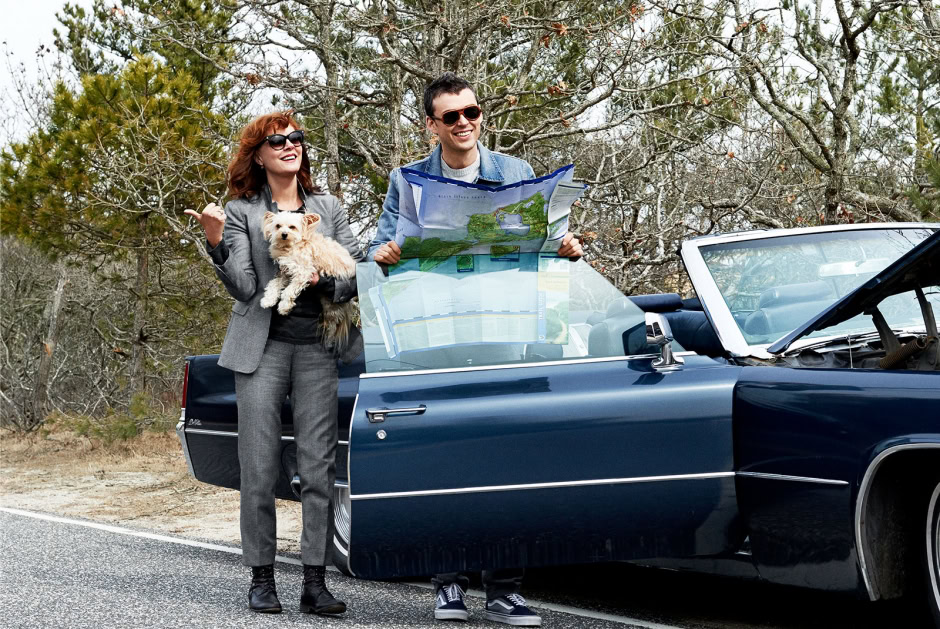 Photo of smiling travelers with a dog stopping by a classic convertible car with open hood and holding a map in a wooded roadside area during daytime.