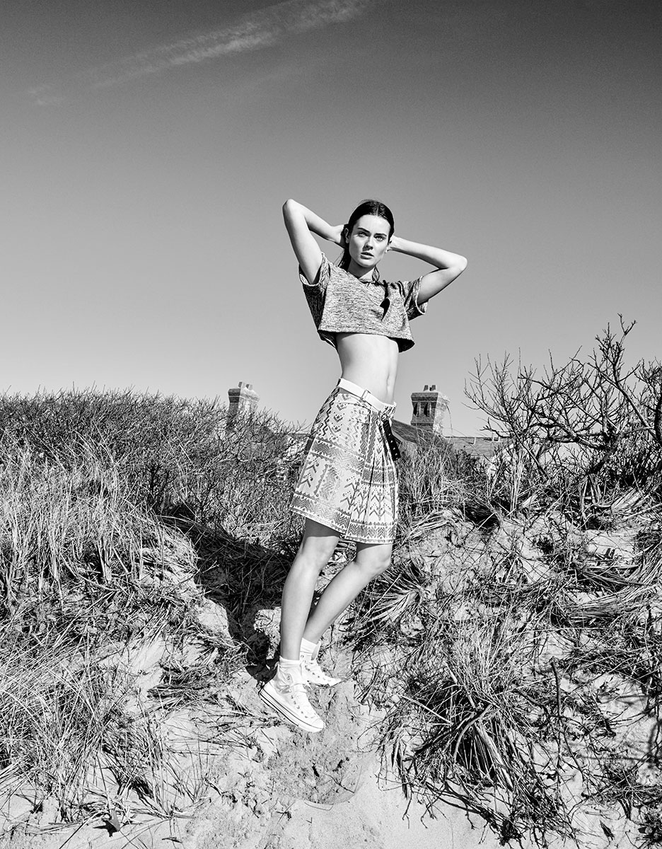 Portrait of a young woman in a casual streetwear outfit posing outdoors on a sandy, grassy area with a clear sky in the background. Black and white photography highlighting fashion and natural scenery.