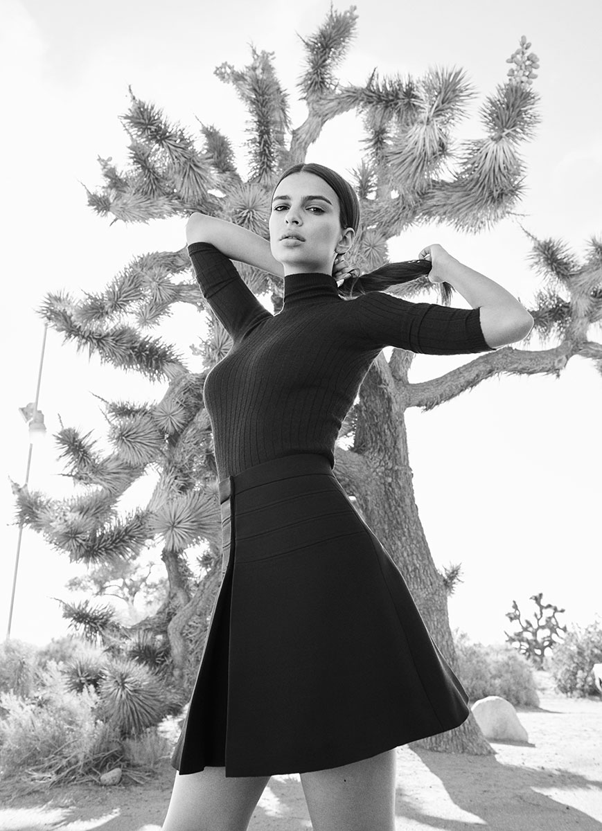 Black and white fashion photography of a woman in a fitted black turtleneck and high-waisted skirt, standing outdoors near a desert tree, emphasizing elegance and timeless style.