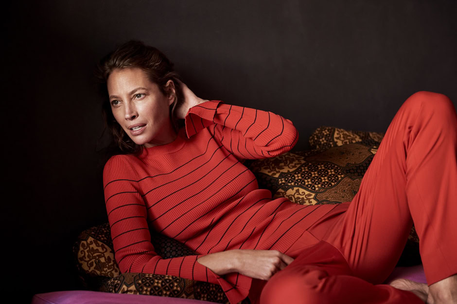 Red striped sweater and matching pants fashion photography by Pamela Hanson, relaxed woman posing on decorative cushions, elegant portrait with dark background, high-quality editorial fashion shot.