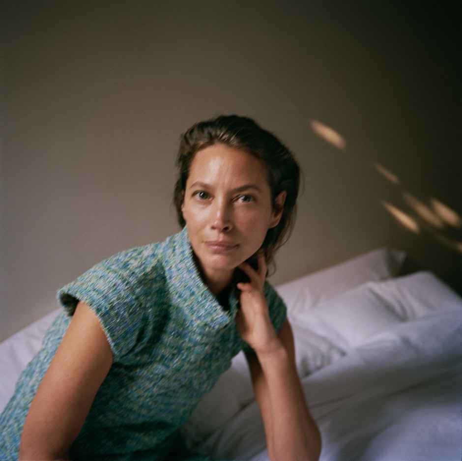 Elegant woman in a textured blue-green sleeveless top sitting on a bed, natural lighting, minimalistic background, artistic portrait photography, sophisticated mood.