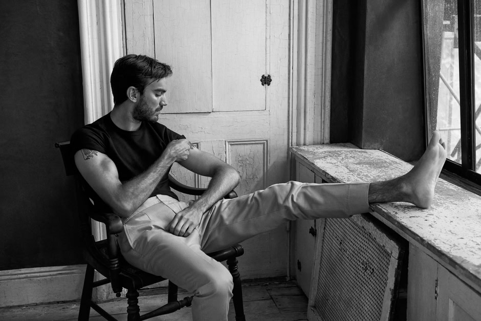 Men sitting by the window with one leg propped up, in a candid black-and-white shot showcasing contemporary portrait photography.
