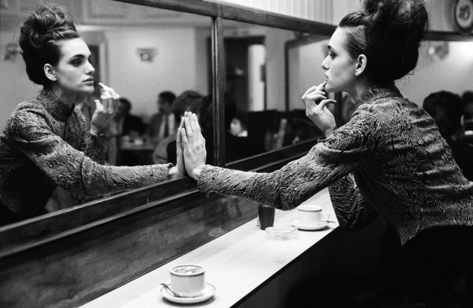 Elegant woman in vintage patterned jacket looking at herself in mirror at café, black and white photo.