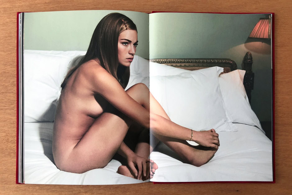 Elegant nude portrait of a young woman with long brown hair sitting on a bed in a luxurious bedroom, showcasing artistic photography by Pamela Hanson.