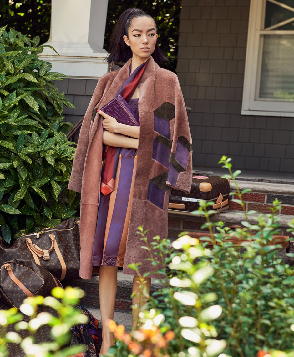 A woman standing on her house steps with colorful travel bags, wearing a statement coat, showcasing upscale fashion photography.