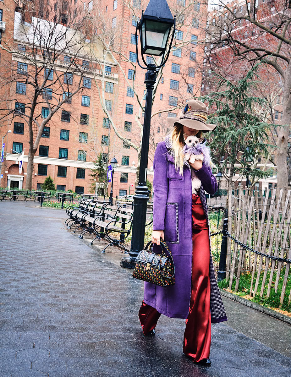 Elegant woman walking in city park holding small dog, stylish purple coat, red satin pants, trendy wide-brim hat, sophisticated handbag, urban photography, fashion portrait, city lifestyle, Pamela Hanson Photography.