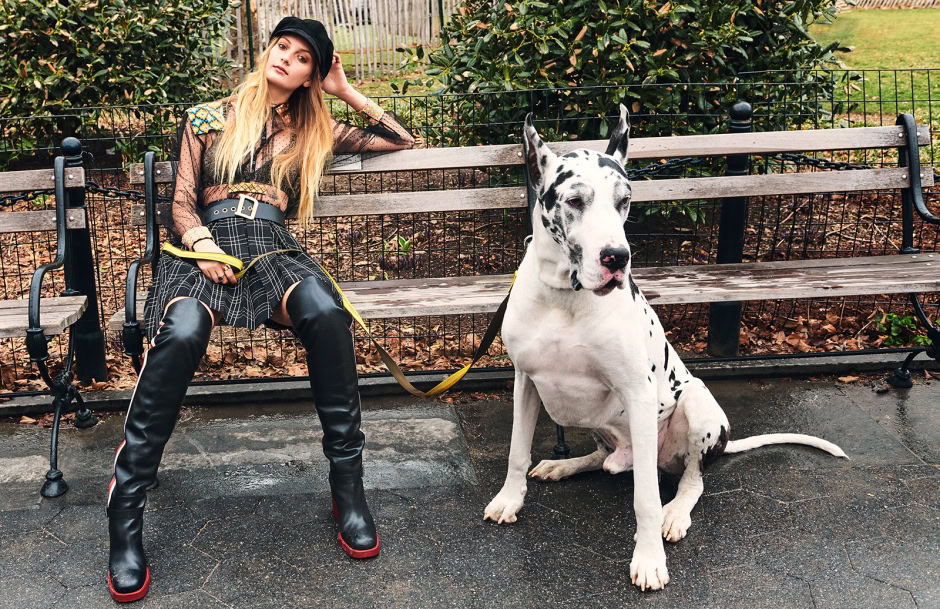 Elegant young woman sitting on a park bench with a Great Dane dog; fashion-forward style, urban outdoor setting, stylish outfit, and trendy accessories.