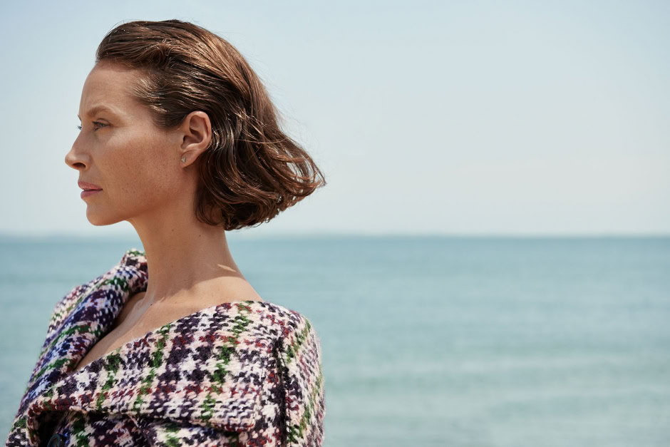 Elegant woman with short brown hair standing by the ocean in a stylish multicolored patterned coat, capturing a serene seaside moment, professional fashion and lifestyle photography by Pamela Hanson.