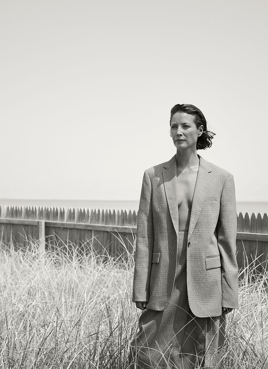 Elegant black and white portrait of a woman in a tailored blazer and trousers, standing outdoors in tall grass with a wooden fence in the background, showcasing professional fashion photography.