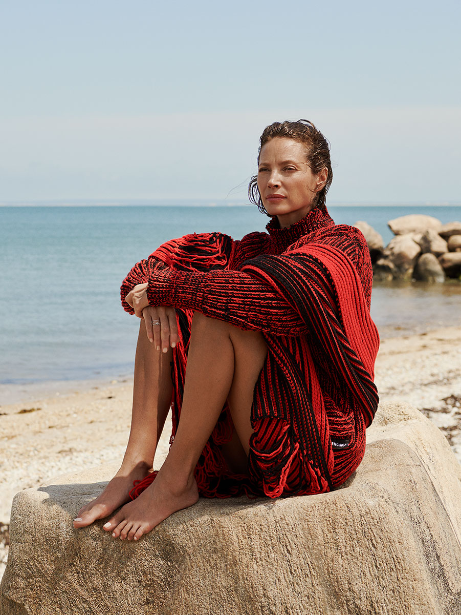Vibrant red and black knit sweater worn by woman sitting on a large rock at the beach with ocean background, capturing fashion photography by Pamela Hanson.