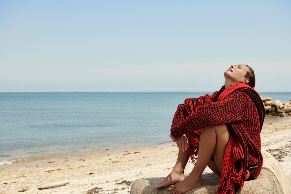 Vibrant red Balenciaga knit blanket with fringe detail draped over a seated woman on a sandy beach by the ocean, enjoying sunny weather and ocean views.
