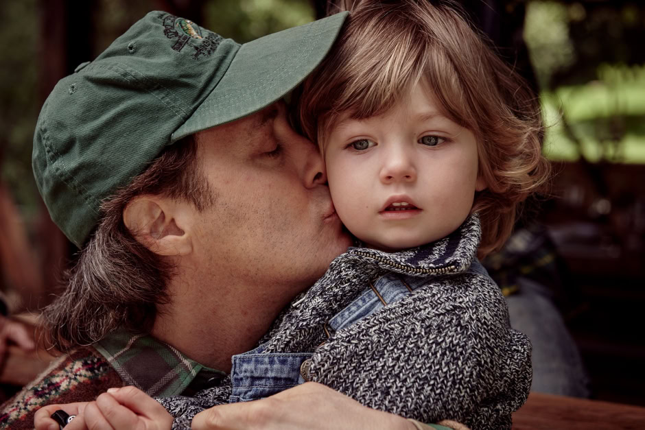 A man wearing a green cap kisses a young boy with curly brown hair, capturing an intimate, candid moment outdoors with natural light.