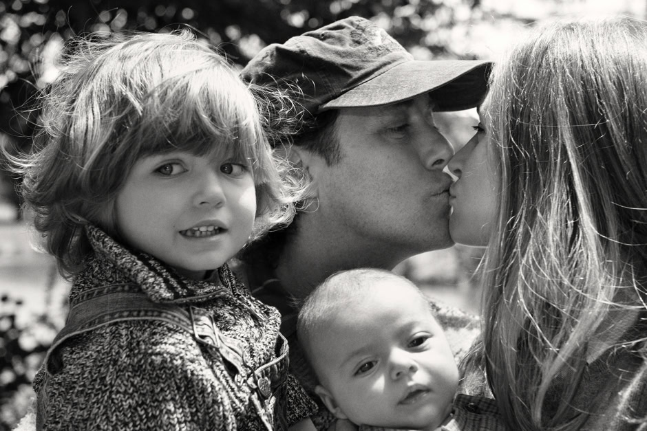 Family portrait capturing a father sharing a tender kiss with his daughter, with two young children observing peacefully; black and white photography emphasizing emotion and intimacy.