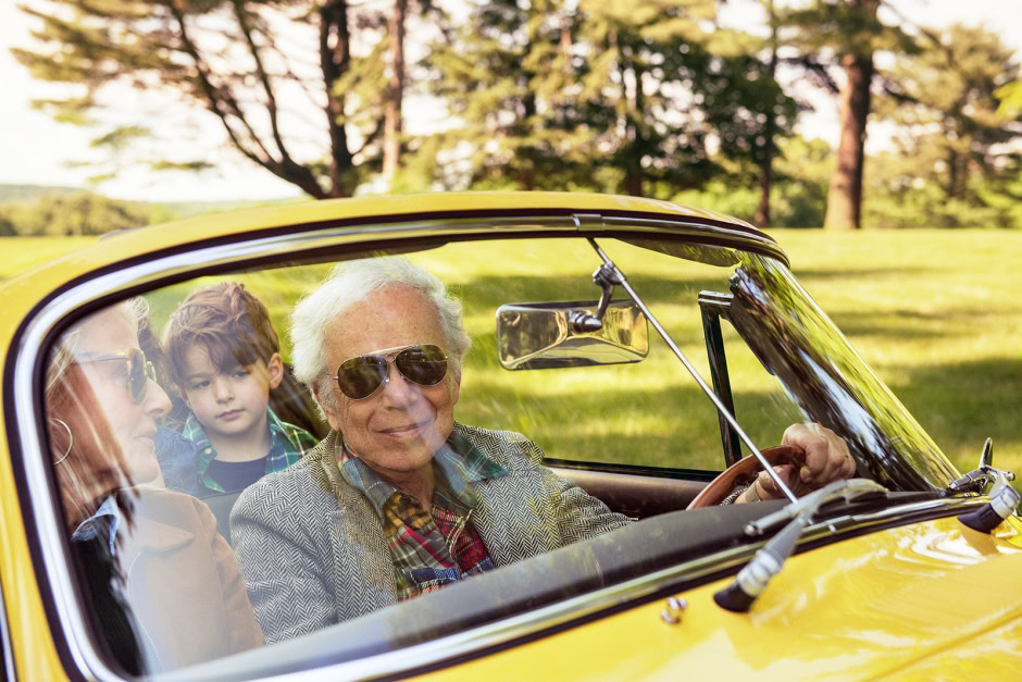 Vintage yellow convertible car with elderly man wearing sunglasses, woman, and young boy enjoying a sunny day in a park, scenic background, classic car photography, lifestyle portrait, outdoor family photoshoot.