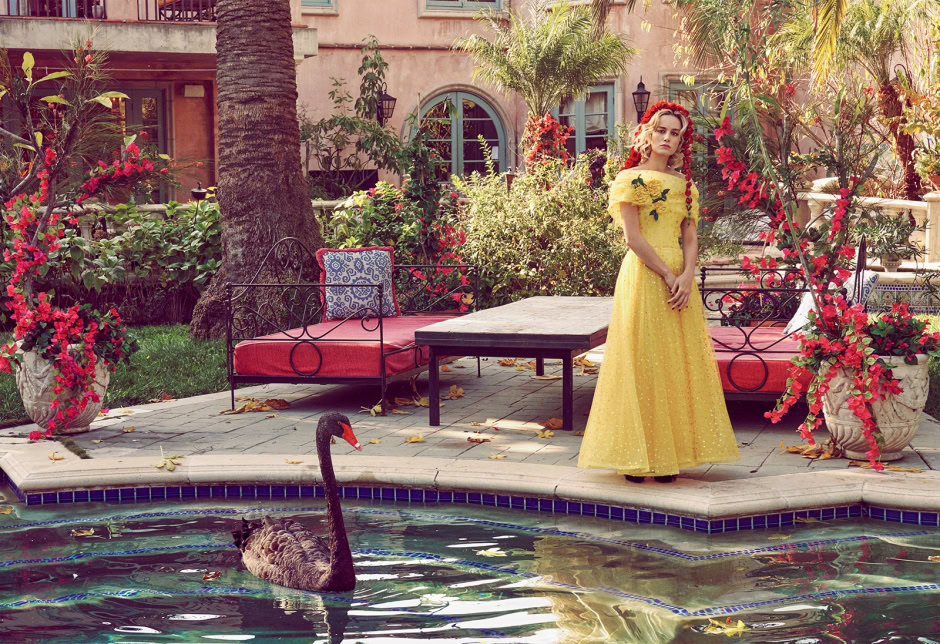 Elegant woman in a yellow dress standing poolside with lush tropical plants and vibrant flowers, capturing high fashion and lifestyle photography in a luxurious outdoor garden setting.