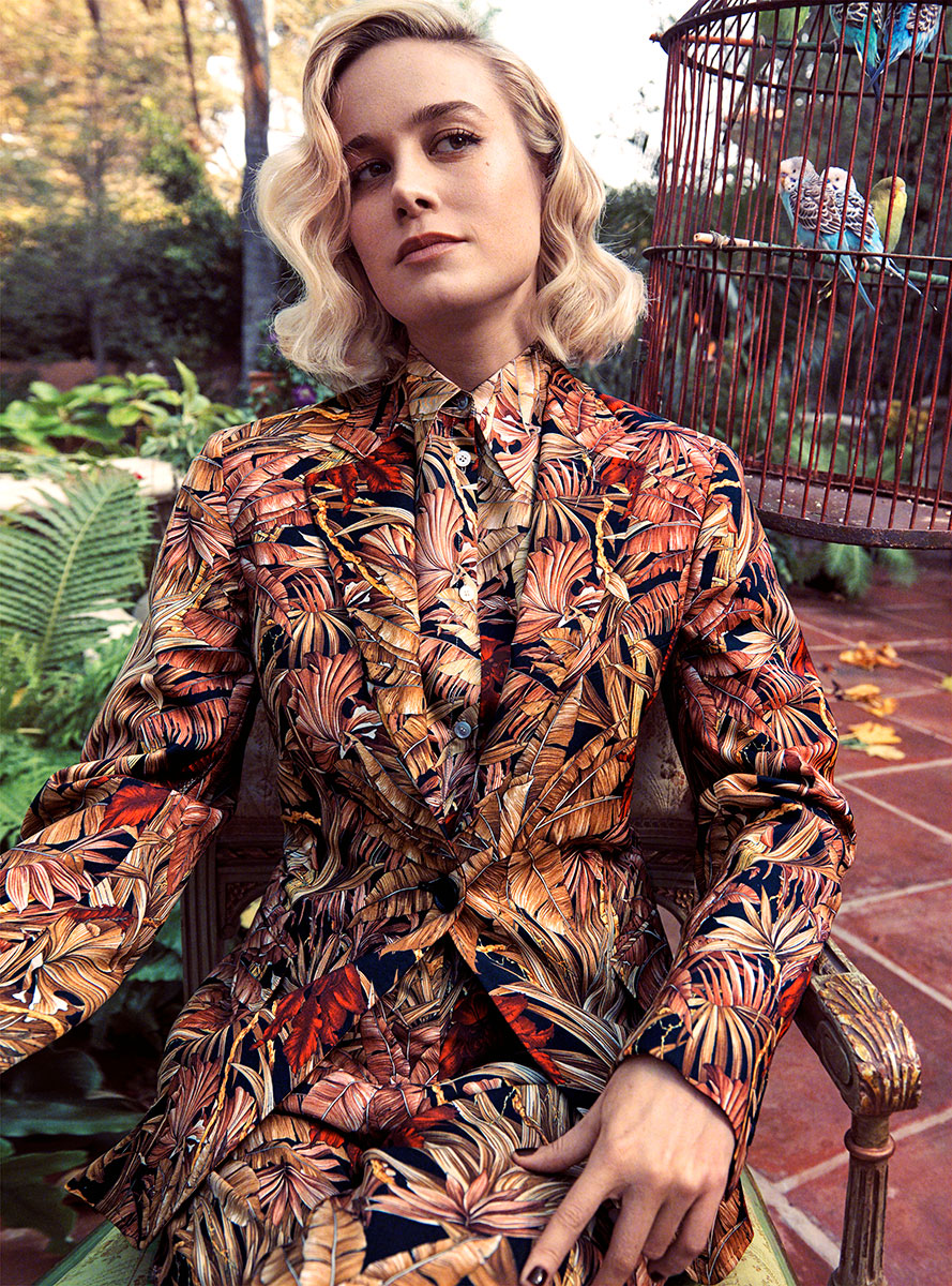 Flowered blazer and matching pants outfit worn by a woman with blonde hair sitting on a garden chair outdoors with colorful birdcage and birds in it, natural greenery background, fashion photography by Pamela Hanson.