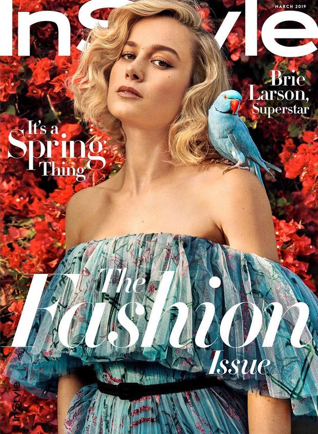 Stunning portrait of a woman with blonde wavy hair wearing a colorful off-shoulder dress, featuring a bright blue bird perched on her shoulder against a vibrant red floral background.