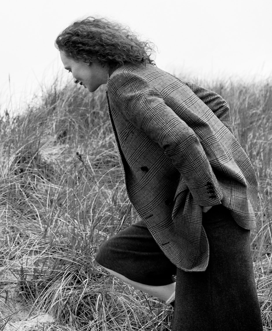 Women's fashion photography, black and white image of woman in plaid blazer and coat in a grassy field, high fashion portrait, timeless style photography, outdoor fashion shoot, professional fashion model, natural environment, artistic fashion photography, Pamela Hanson Photography relevance.