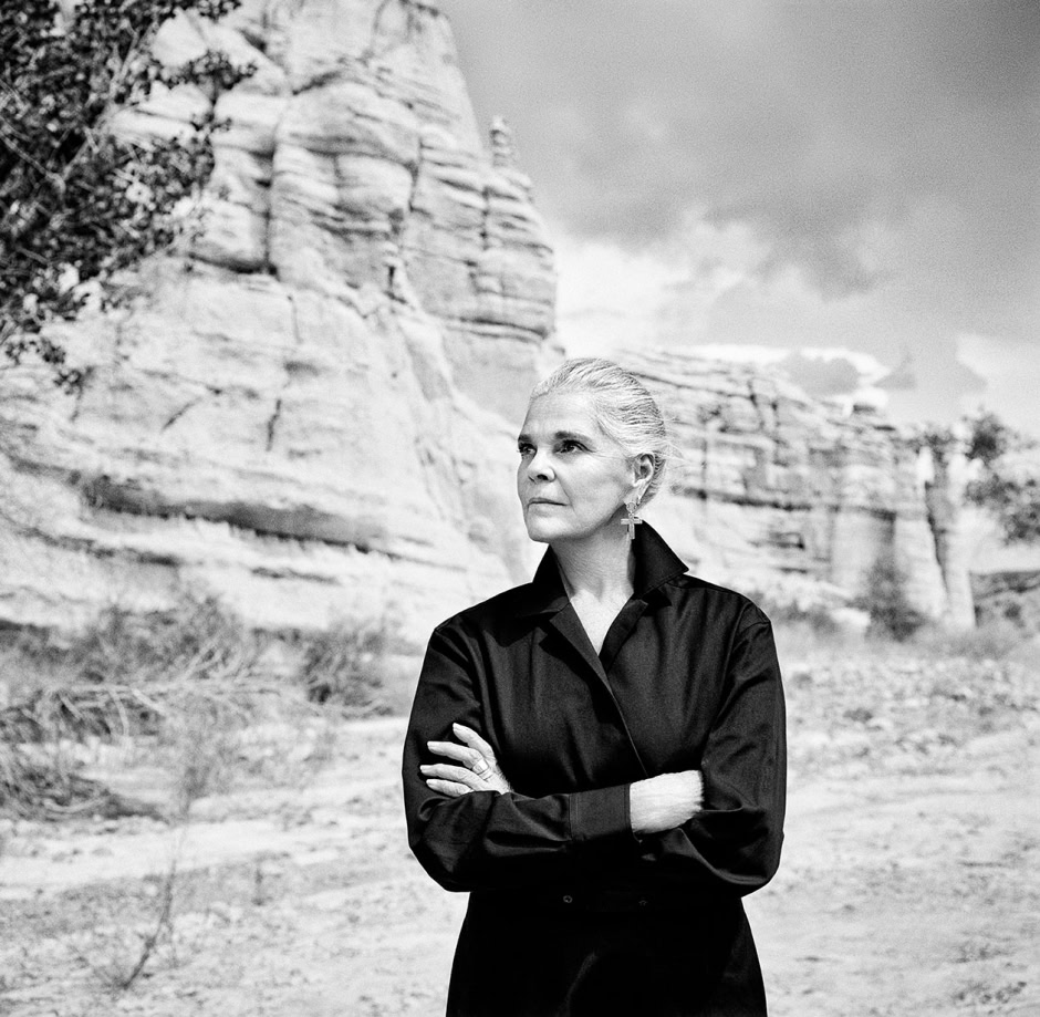 Elegant woman in black shirt with crossed arms standing outdoors near rock formations, capturing a moment of reflection; portrait by Pamela Hanson Photography emphasizing fashion and natural scenery.