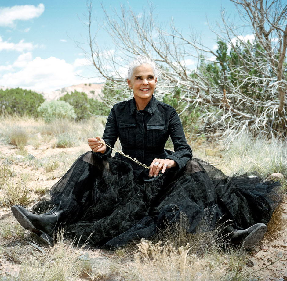 Elegant senior woman outdoor portrait with silver hair, black fashionable dress, sitting on desert terrain surrounded by shrubs and drought-tolerant trees, under a sunny blue sky.