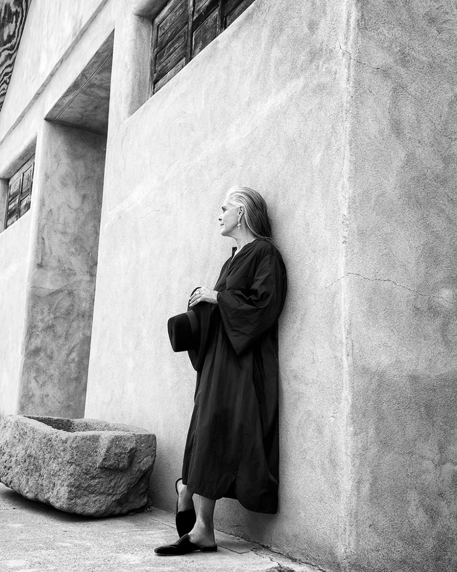 Elegant black and white portrait of a middle-aged woman standing against a textured concrete wall, holding a hat and wearing stylish clothing, showcasing sophisticated fashion photography.