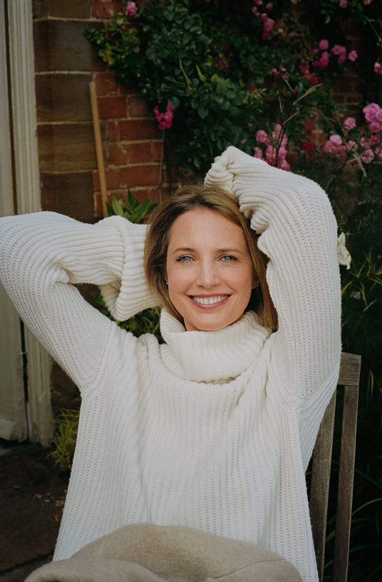 Elegant portrait of a woman in a cozy white knit sweater, smiling outdoors amidst vibrant pink and green garden foliage, showcasing natural beauty and relaxed outdoor photography.