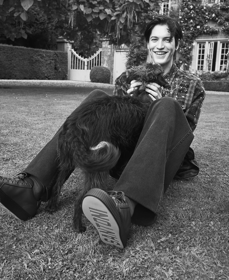 Young man smiling playing with puppies in a garden, black and white photo, contemporary portrait of youthful joy and pet companionship, outdoor setting with lush greenery and elegant house background, lifestyle photography, candid moment, relaxed atmosphere, authentic connection between person and dog, professional photography shot.