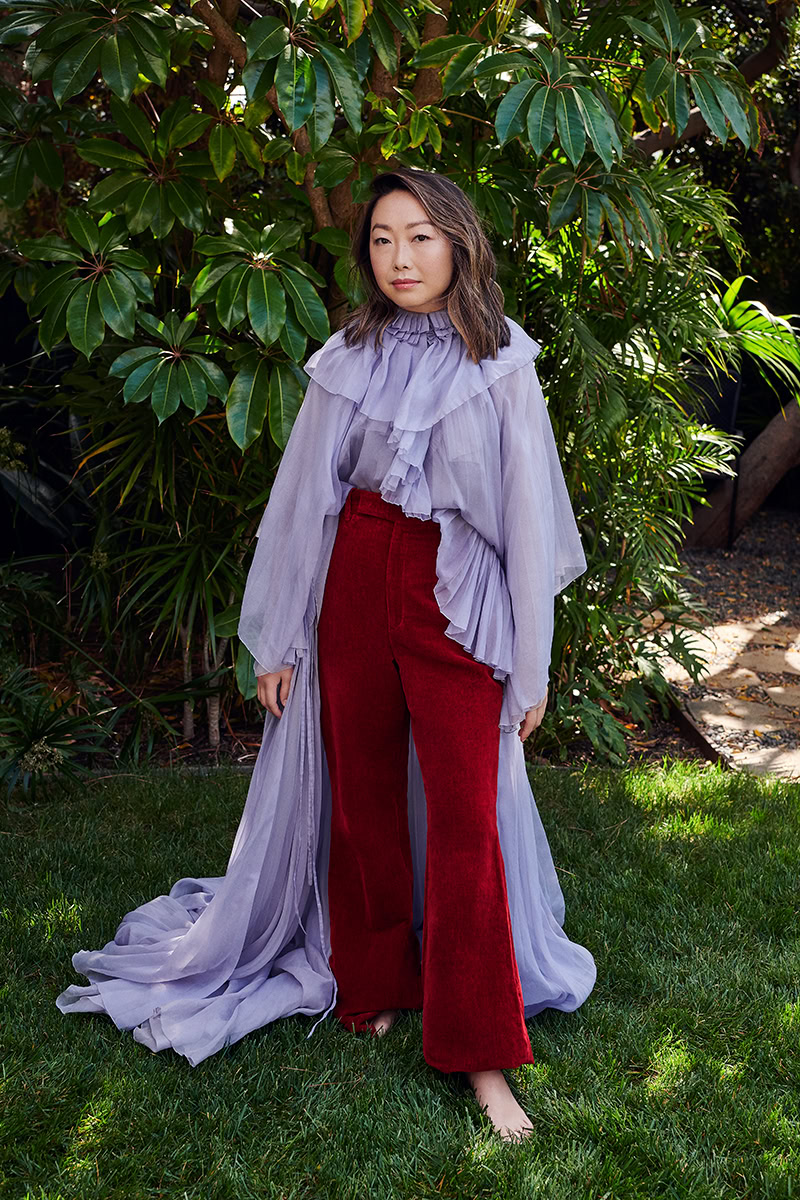 Woman standing in lush garden with large green leaves and sunlight, wearing purple ruffled blouse and red corduroy pants, showcasing sophisticated fashion photography by Pamela Hanson.
