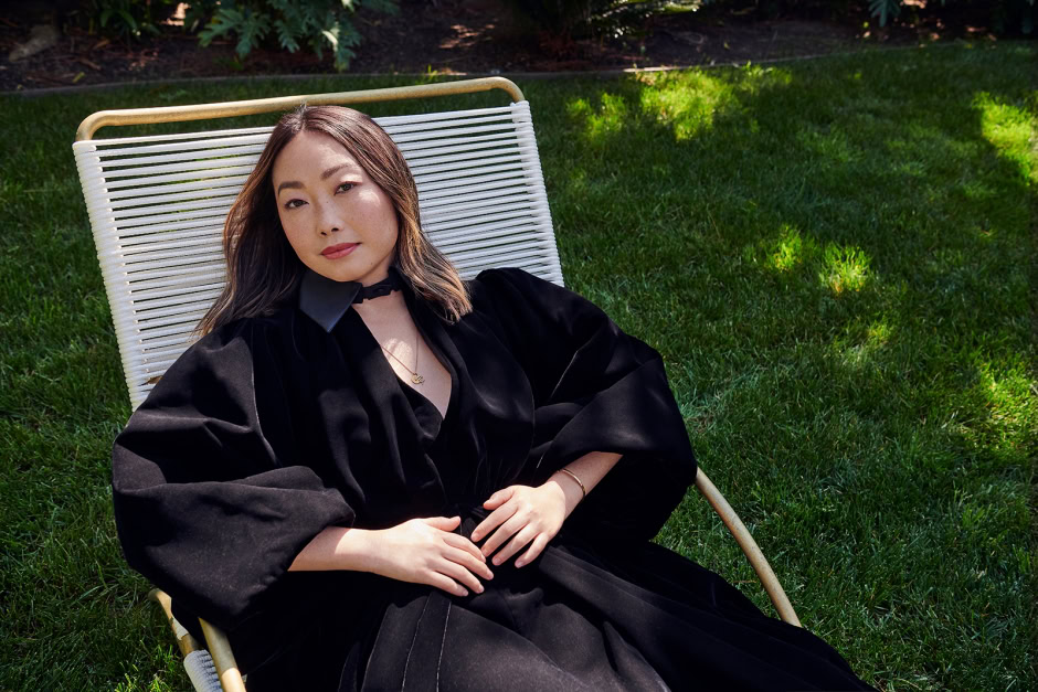 Elegant portrait of a woman in a black dress relaxing outdoors on a white lounge chair with lush green grass and sunlight in the background.