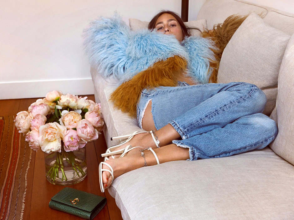 Soft-focus portrait of a woman lounging on a beige sofa, wearing casual distressed jeans and a fluffy blue and brown fur jacket, with a bouquet of pink peonies on a wooden table beside her.