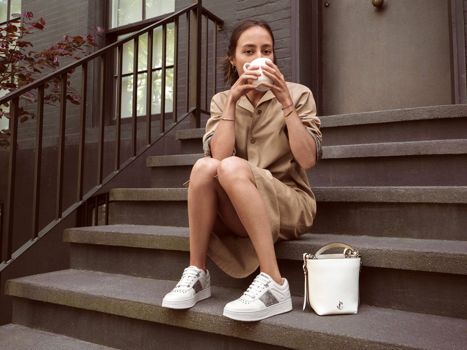 Comfortable woman sitting on outdoor stairs, casually drinking from a mug, wearing a beige dress and white sneakers, with a stylish white handbag beside her, captured in a relaxed lifestyle fashion photography session.