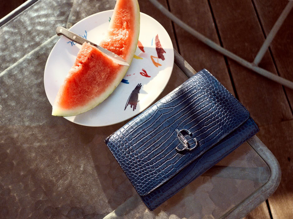 Juicy watermelon slice on a white plate with colorful animal illustrations, placed on a rustic outdoor table alongside a chic blue crocodile-embossed clutch bag with a metallic logo. Perfect summer day photo.