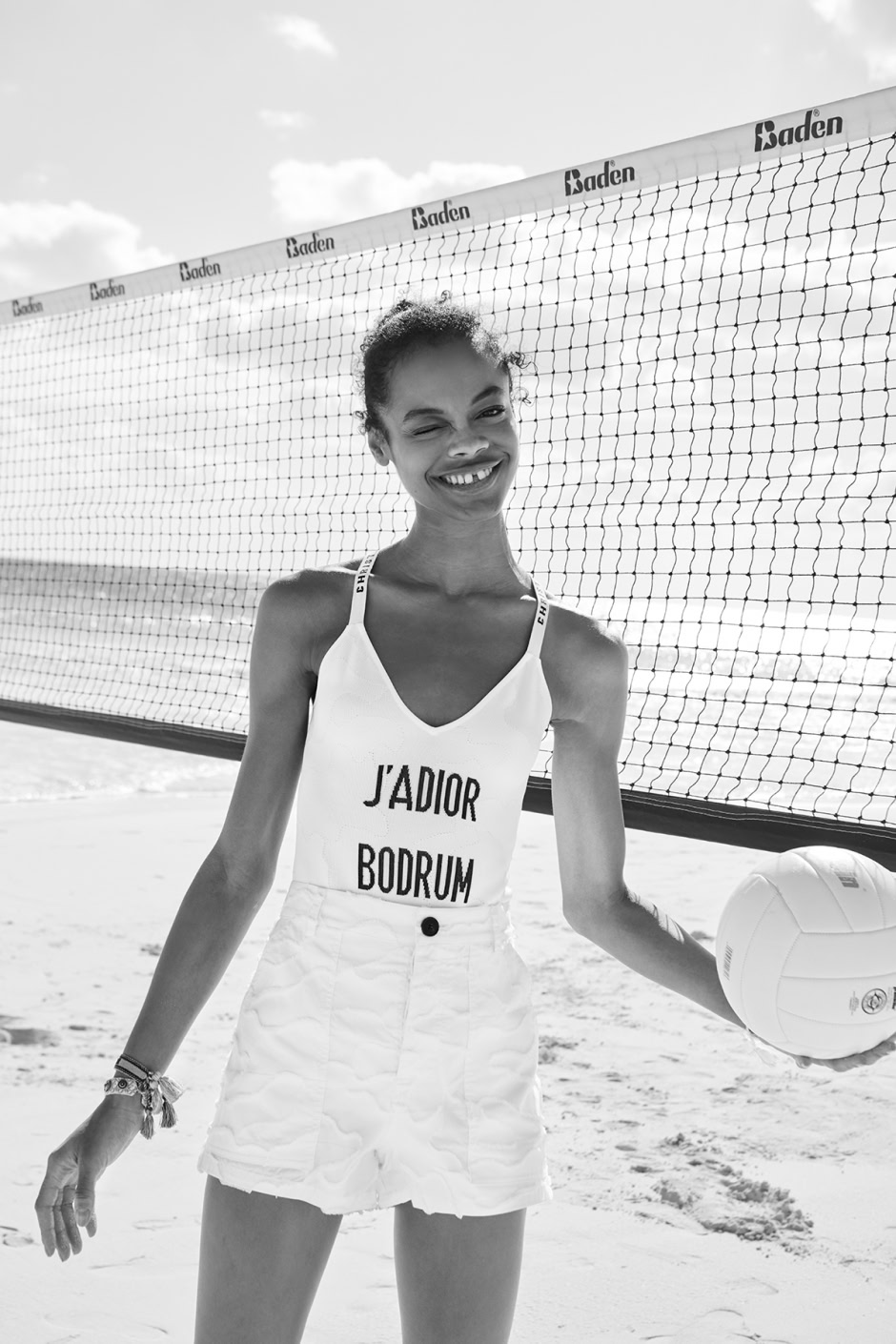 Stunning black and white photo of a young female beach volleyball player smiling on the sandy court, holding a volleyball, with a net and ocean backdrop.