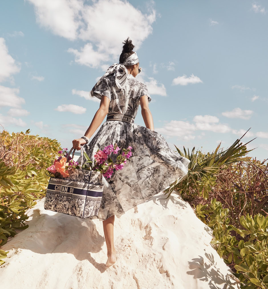 Luxurious fashion model wearing floral Christian Dior dress with matching accessories, carrying a designer tote with vibrant flowers, on a sunlit sandy dune with lush green foliage and blue sky background.